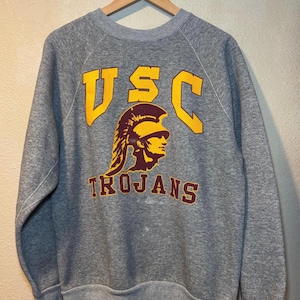 May include: A heather grey crewneck sweatshirt featuring the yellow text USC above a graphic of a maroon and yellow Trojan helmeted profile, with the word TROJANS printed in maroon below the graphic, all displayed on a wooden hanger.