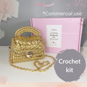Könnte beinhalten: Eine goldene Häkeltasche mit passendem Kettenträger und Henkel. Die Tasche hat eine rechteckige Form mit einem Verschluss. Eine rosa Box mit dem Text "Crochet kit Lili" befindet sich im Hintergrund.