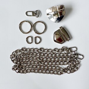 May include: A set of silver-toned metal hardware for a bag or purse. The set includes two D-rings, two swivel clasps, two lobster clasps, a chain, and two metal pieces with a shield shape.