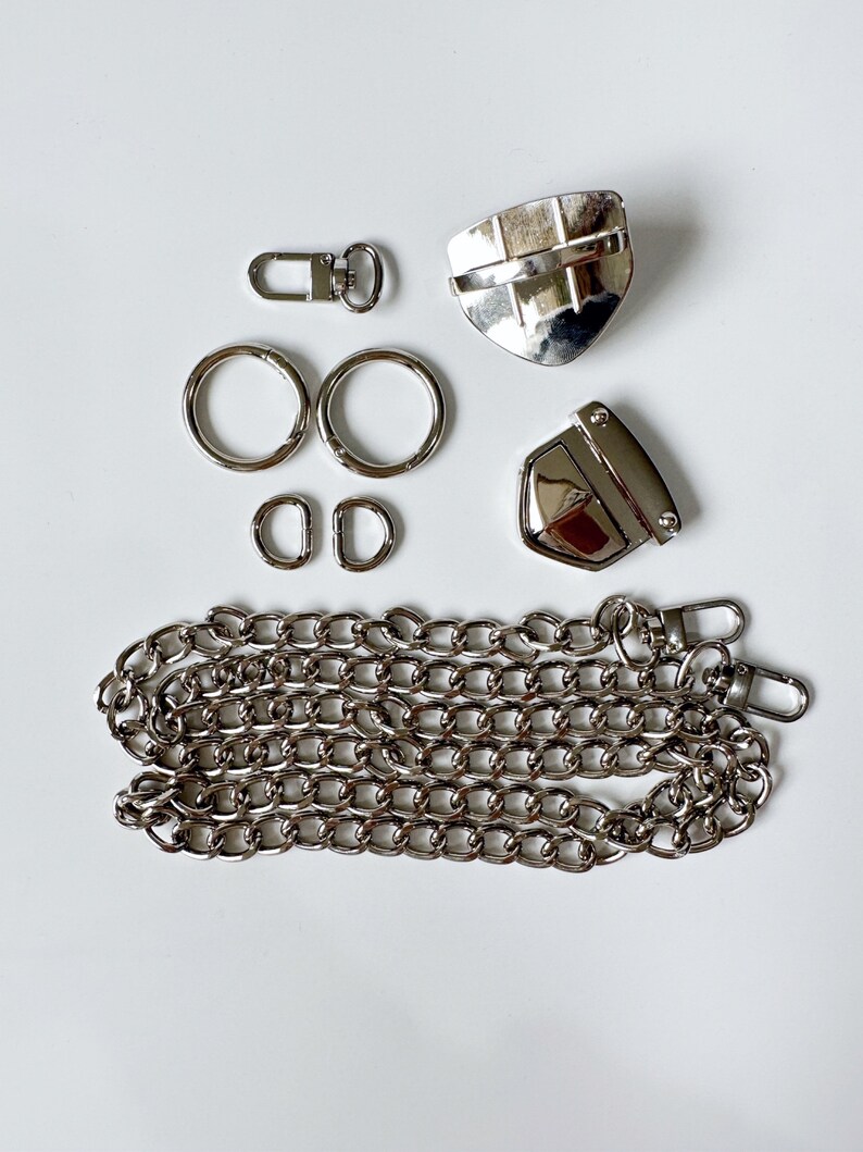 May include: A set of silver-toned metal hardware for a bag or purse. The set includes two D-rings, two swivel clasps, two lobster clasps, a chain, and two metal pieces with a shield shape.