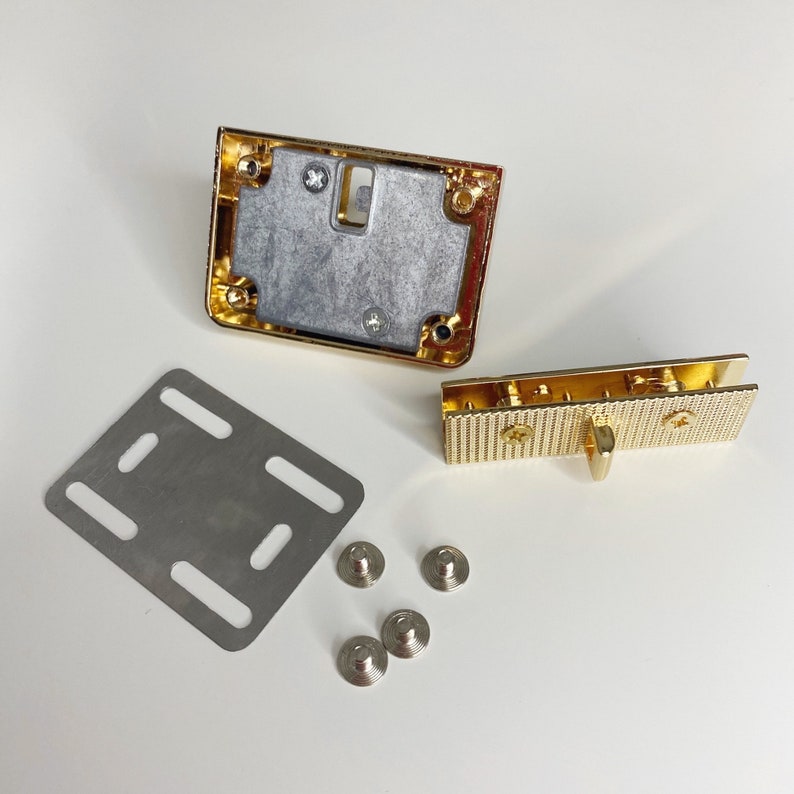 May include: Gold-toned metal hardware set for a bag or purse. The set includes a rectangular frame, a smaller rectangular piece with a latch, and four small metal studs.