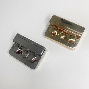 May include: Two metal clasps with a rectangular shape and three round buttons. One clasp is silver and the other is gold.