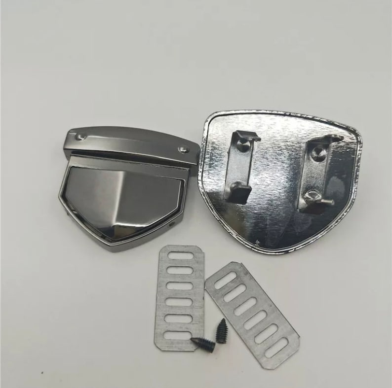 May include: A set of silver metal belt buckle hardware. The set includes two shield-shaped pieces, two rectangular pieces with holes, and a small silver screw.