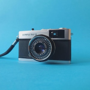 Op de afbeelding: Olympus Trip 35 camera met een zwart en zilverkleurig lichaam en een lensdop. De camera heeft een zwarte band en staat tegen een effen blauwe achtergrond. De lens is gemarkeerd met "Made in Japan".