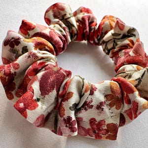 May include: A floral scrunchie with a cream background and red, orange, and burgundy flower pattern. The fabric is gathered and sewn into a circular shape, perfect for securing hair in a ponytail or bun.