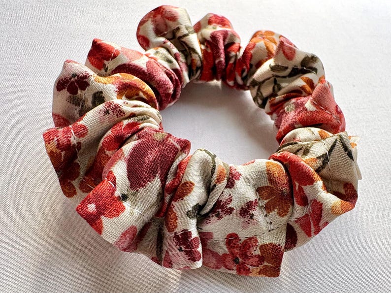 May include: A floral scrunchie with a cream background and red, orange, and burgundy flower pattern. The fabric is gathered and sewn into a circular shape, perfect for securing hair in a ponytail or bun.