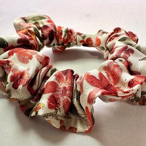 May include: A floral scrunchie with a white background and red and pink flower pattern. The fabric is gathered and elasticized, creating a circular shape. The scrunchie is a hair accessory.
