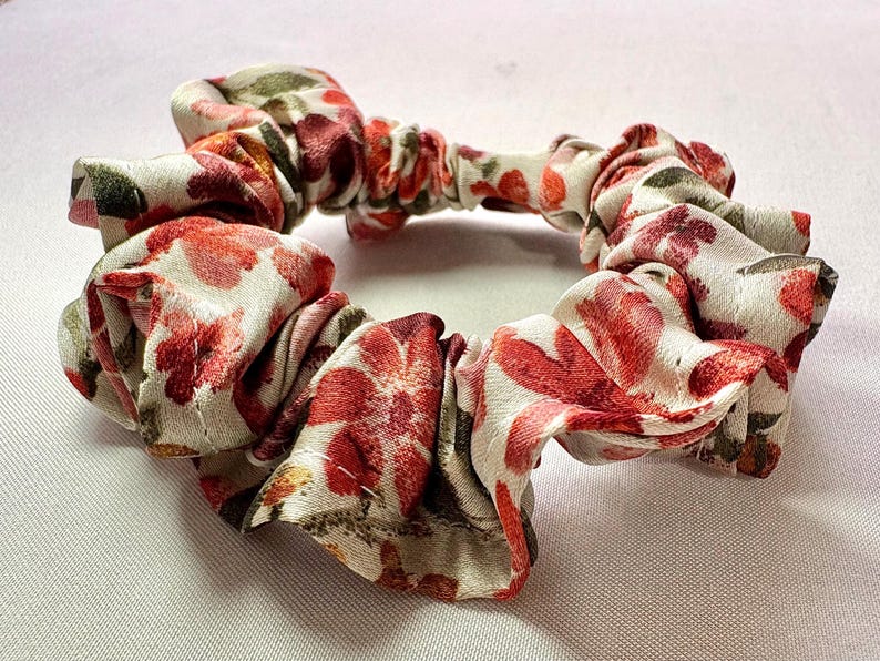 May include: A floral scrunchie with a white background and red and pink flower pattern. The fabric is gathered and elasticized, creating a circular shape. The scrunchie is a hair accessory.