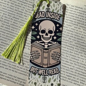 以下が含まれることがあります： 本を読む骸骨が描かれたブックマークで、「DEAD INSIDE BUT WELL READ」の文字が入っています。 ブックマークには緑色のタッセルと、骸骨と葉の装飾模様が付いています。