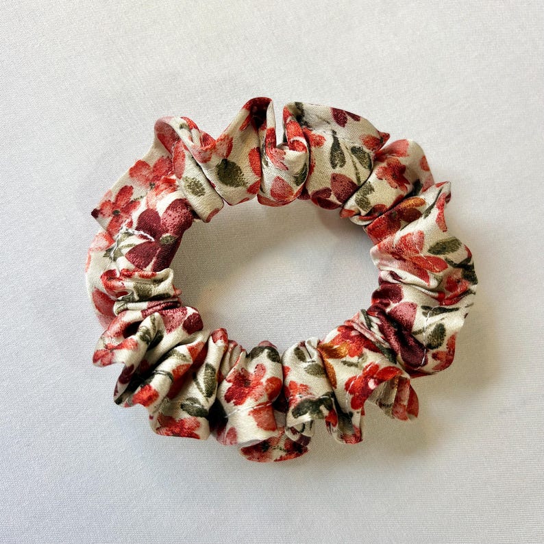 May include: A floral scrunchie with a cream background and red and green flower pattern. The fabric is gathered and ruffled, creating a circular shape. This hair accessory adds a touch of elegance.