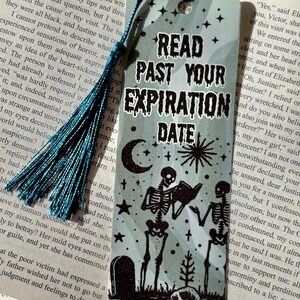 以下が含まれることがあります： ターコイズのタッセルが付いた長方形のブックマーク。「READ PAST YOUR EXPIRATION DATE」というテキストが、本を読む2体の骸骨のグラフィックの上に表示されています。月、星、墓石も描かれています。
