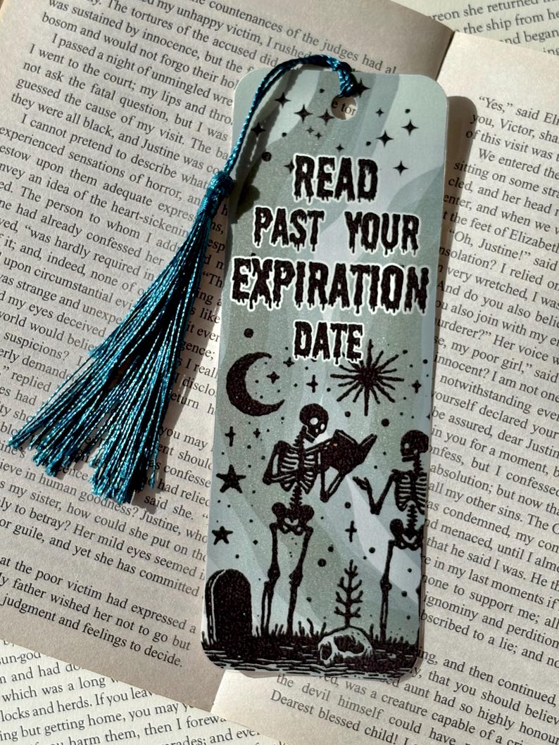 以下が含まれることがあります： ターコイズのタッセルが付いた長方形のブックマーク。「READ PAST YOUR EXPIRATION DATE」というテキストが、本を読む2体の骸骨のグラフィックの上に表示されています。月、星、墓石も描かれています。