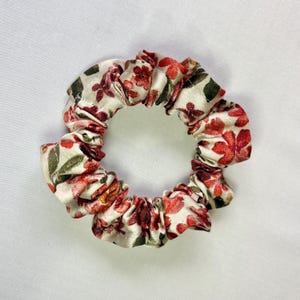 May include: A floral scrunchie featuring a cream background with red and green flower patterns. The fabric is gathered and elasticized, creating a circular shape. This hair accessory is suitable for various hairstyles.