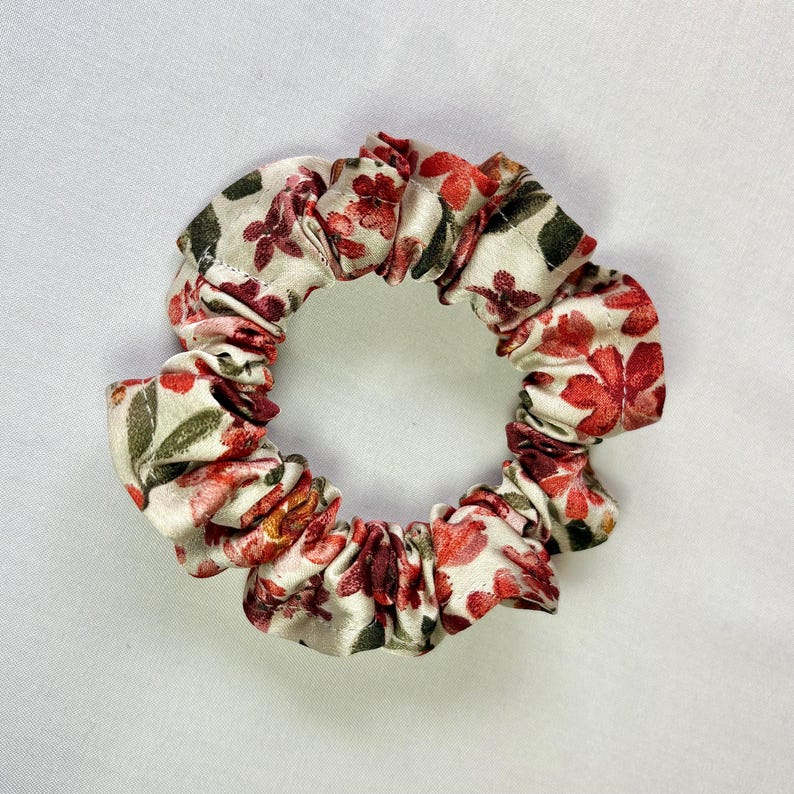 May include: A floral scrunchie featuring a cream background with red and green flower patterns. The fabric is gathered and elasticized, creating a circular shape. This hair accessory is suitable for various hairstyles.