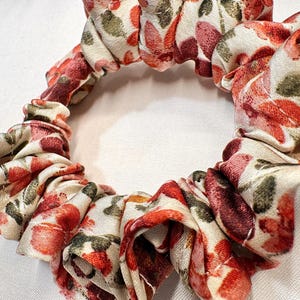 May include: A floral scrunchie with a cream background and red, pink, and green flower pattern. The fabric is gathered and elasticized, creating a ruffled effect. This hair accessory is suitable for various hairstyles.