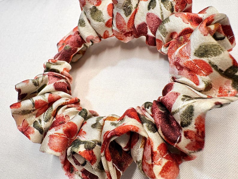 May include: A floral scrunchie with a cream background and red, pink, and green flower pattern. The fabric is gathered and elasticized, creating a ruffled effect. This hair accessory is suitable for various hairstyles.