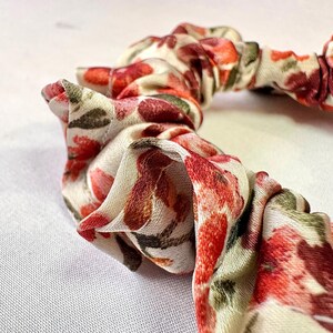May include: Close-up of a floral scrunchie. The fabric features a pattern of red, pink, and brown flowers with green leaves on a cream background. The scrunchie is gathered and has a soft, textured appearance.