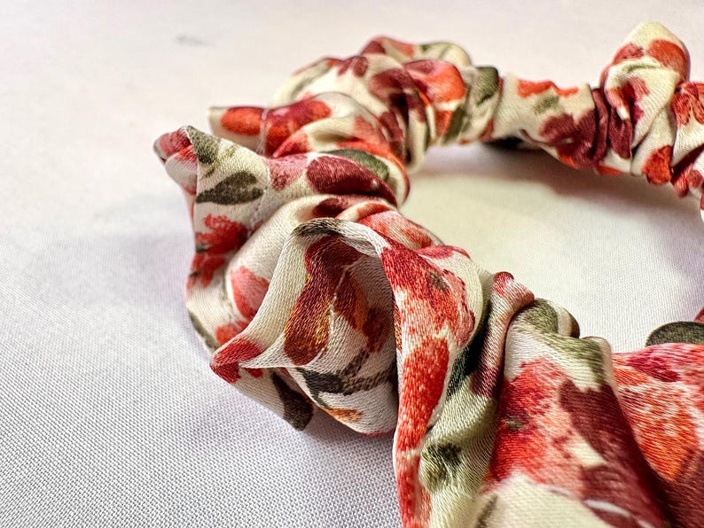 May include: Close-up of a floral scrunchie. The fabric features a pattern of red, pink, and brown flowers with green leaves on a cream background. The scrunchie is gathered and has a soft, textured appearance.
