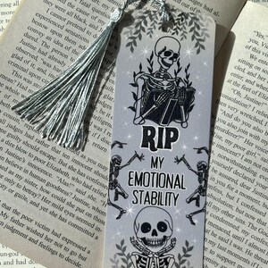以下が含まれることがあります： グレーの背景に、本を読む骸骨が描かれたブックマークで、「RIP MY EMOTIONAL STABILITY」というテキストが入っています。 ブックマークにはシルバーのタッセルが付いています。