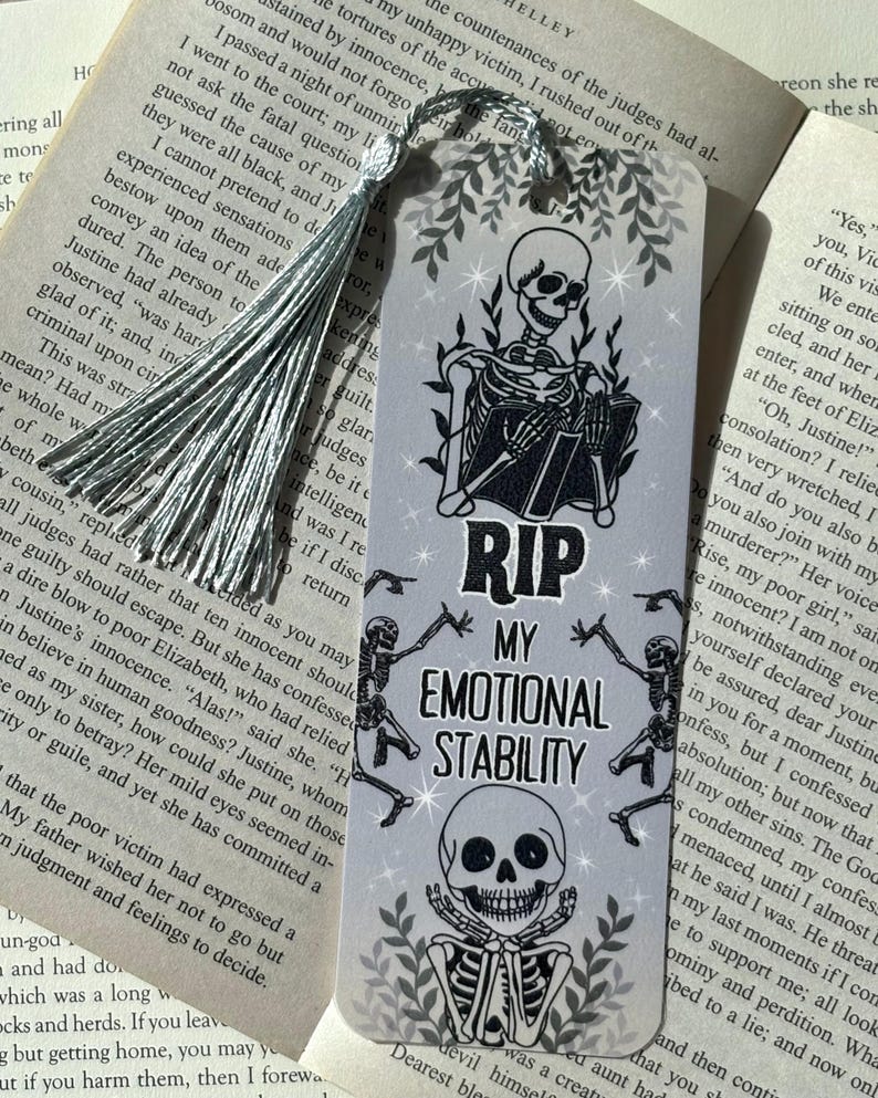 以下が含まれることがあります： グレーの背景に、本を読む骸骨が描かれたブックマークで、「RIP MY EMOTIONAL STABILITY」というテキストが入っています。 ブックマークにはシルバーのタッセルが付いています。