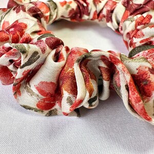 May include: A floral scrunchie with a white background and a pattern of red, pink, and green flowers. The fabric is gathered and twisted, creating a textured appearance. This hair accessory is suitable for various hairstyles.