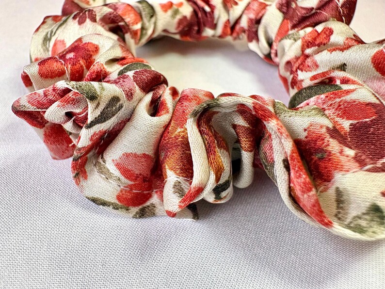 May include: A floral scrunchie with a white background and a pattern of red, pink, and green flowers. The fabric is gathered and twisted, creating a textured appearance. This hair accessory is suitable for various hairstyles.