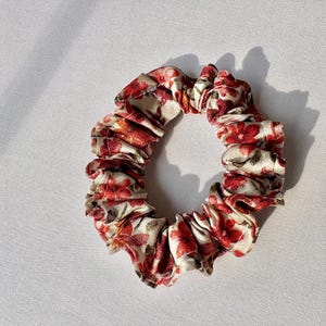 May include: A floral scrunchie featuring a pattern of red flowers and green leaves on a cream background. The fabric is gathered, creating a ruffled appearance. This hair accessory is designed to hold hair in place.
