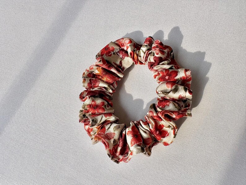 May include: A floral scrunchie featuring a pattern of red flowers and green leaves on a cream background. The fabric is gathered, creating a ruffled appearance. This hair accessory is designed to hold hair in place.