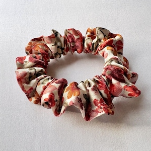 May include: A floral scrunchie featuring a cream background with red, orange, and pink flower patterns. The fabric is gathered and elasticized, creating a ruffled appearance. This hair accessory is suitable for various hairstyles.