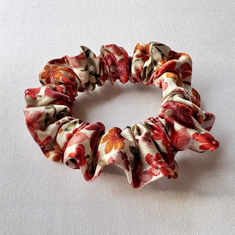 May include: A floral scrunchie featuring a cream background with red, orange, and pink flower patterns. The fabric is gathered and elasticized, creating a ruffled appearance. This hair accessory is suitable for various hairstyles.
