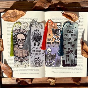 以下が含まれることがあります： 骸骨のイラストと「DEAD INSIDE」や「RIP MY EMOTIONAL STABILITY」などのユーモラスなフレーズが描かれた4つの装飾的なブックマーク。各ブックマークには異なる色のタッセルが付いており、開いた本の上に置かれています。