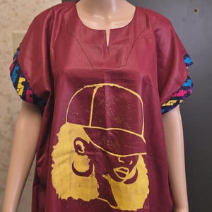 May include: A maroon tunic dress featuring a yellow graphic of a person wearing a cap. The dress has short sleeves with colorful patterned cuffs and a notched neckline. The dress is displayed on a mannequin.