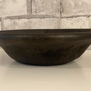 May include: A dark brown, shallow bowl with a wide rim. The bowl has a rounded shape and sits on a white surface. The background is a light grey brick wall.