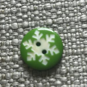 May include: A round, green button featuring a white snowflake design centered on the surface. The button has two small, circular holes positioned in the middle of the white snowflake pattern, allowing for attachment to fabric or other materials.