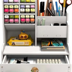 May include: White desktop organizer with multiple compartments for storing pens, pencils, markers, and other office supplies. It has a drawer for storing smaller items and a top shelf for displaying items.