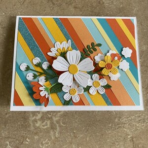 Puede incluir: Tarjeta de felicitaci&oacute;n hecha a mano con un dise&ntilde;o floral. La tarjeta tiene un fondo de rayas diagonales en naranja, amarillo y turquesa. Flores de papel blancas y amarillas con hojas verdes est&aacute;n adheridas al frente.