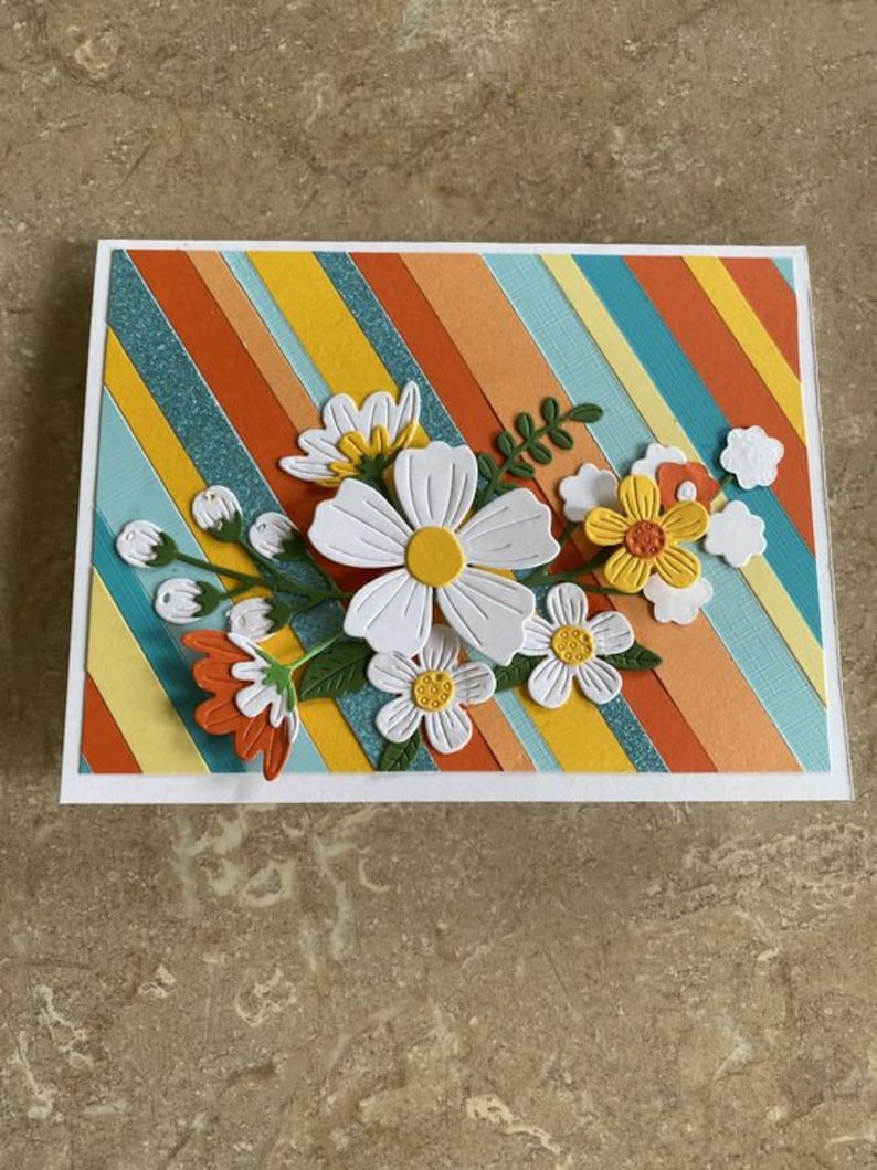 Puede incluir: Tarjeta de felicitaci&oacute;n hecha a mano con un dise&ntilde;o floral. La tarjeta tiene un fondo de rayas diagonales en naranja, amarillo y turquesa. Flores de papel blancas y amarillas con hojas verdes est&aacute;n adheridas al frente.