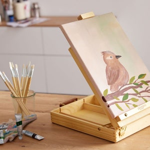 Könnte beinhalten: Ein Holzstaffelei mit einer Leinwand, auf der ein gemalter Vogel auf einem Ast zu sehen ist. Die Staffelei hat ein Ablagefach für Malutensilien. Die Leinwand ist etwa 30 cm x 40 cm groß.