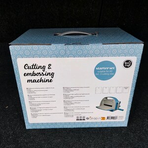 Puede incluir: Una caja con estampado azul y blanco con asa, etiquetada "Cutting & embossing machine". La caja contiene un kit de inicio adecuado para papel A5, que incluye 3 troqueles. Se ve una m&aacute;quina de corte azul y blanca.