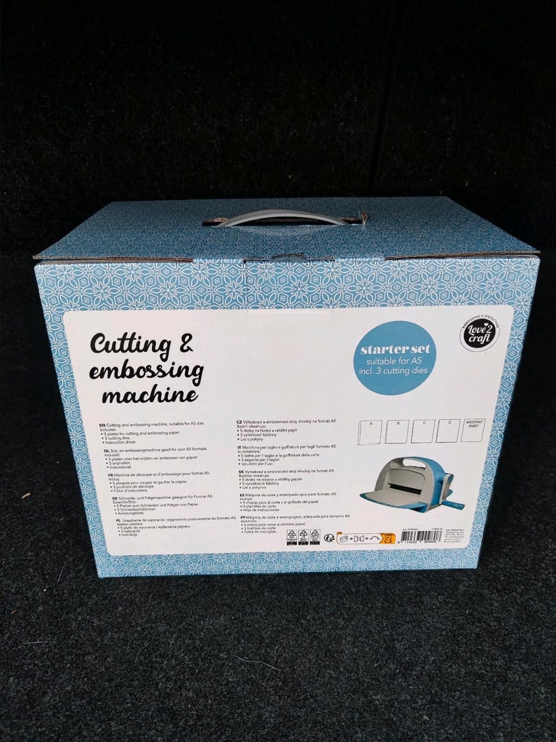Puede incluir: Una caja con estampado azul y blanco con asa, etiquetada "Cutting & embossing machine". La caja contiene un kit de inicio adecuado para papel A5, que incluye 3 troqueles. Se ve una m&aacute;quina de corte azul y blanca.
