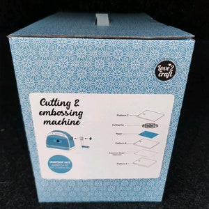 Puede incluir: Una caja azul y blanca estampada para una m&aacute;quina de cortar y grabar en relieve. La caja tiene el texto "Cutting & embossing machine" y "Love 2 Craft". La caja tambi&eacute;n muestra la m&aacute;quina y sus componentes.