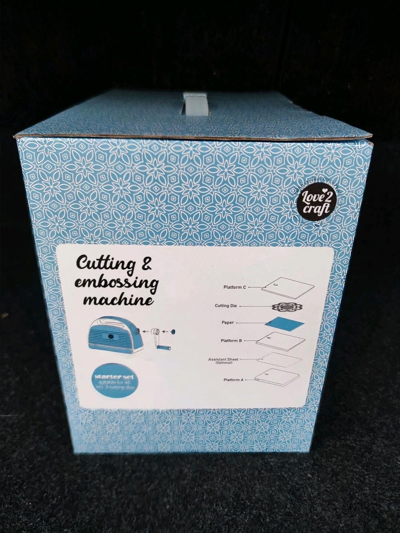 Puede incluir: Una caja azul y blanca estampada para una m&aacute;quina de cortar y grabar en relieve. La caja tiene el texto "Cutting & embossing machine" y "Love 2 Craft". La caja tambi&eacute;n muestra la m&aacute;quina y sus componentes.