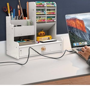 May include: White desktop organizer with a drawer and multiple compartments for storing pens, pencils, markers, and other office supplies. The organizer has a small drawer with a gold knob and is sitting on a white desk with a laptop.