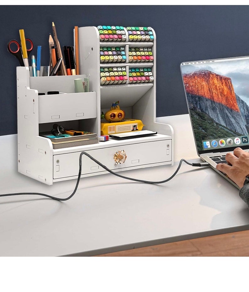 May include: White desktop organizer with a drawer and multiple compartments for storing pens, pencils, markers, and other office supplies. The organizer has a small drawer with a gold knob and is sitting on a white desk with a laptop.