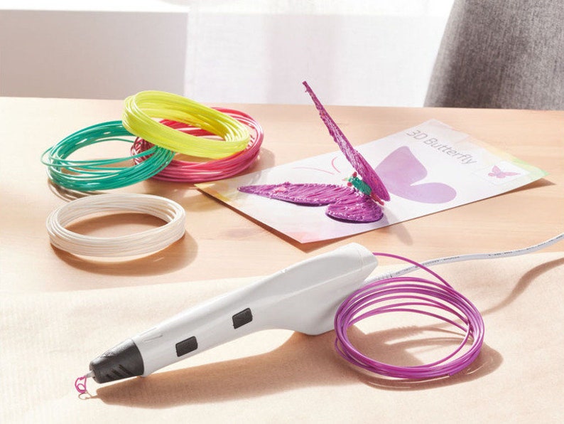 Pu&ograve; includere: Una penna per stampa 3D bianca con punta e pulsanti neri, accanto a una bobina di filamento viola. Sono presenti anche bobine di filamento colorate in verde, giallo, rosa e bianco. Una farfalla 3D viola parzialmente completata &egrave; su una carta.