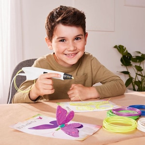 Pu&ograve; includere: Una persona tiene in mano una penna 3D bianca, con filamento giallo che ne esce. Una farfalla viola stampata in 3D &egrave; su un foglio di carta. Sul tavolo ci sono bobine di filamento colorato, tra cui giallo, verde, blu e bianco.