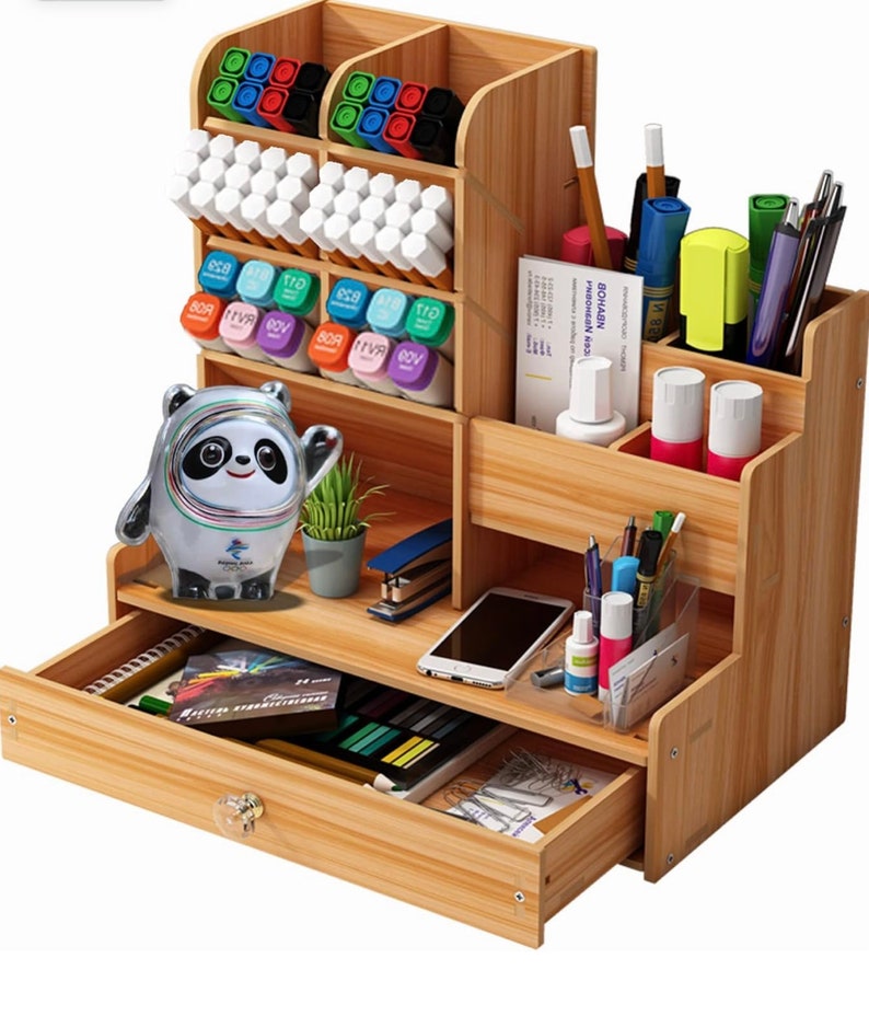 May include: A wooden desk organizer with multiple compartments for pens, pencils, markers, and other office supplies. It has a drawer for storing larger items and a small shelf for a phone or other devices. The organizer is made of natural wood and has a light brown finish.