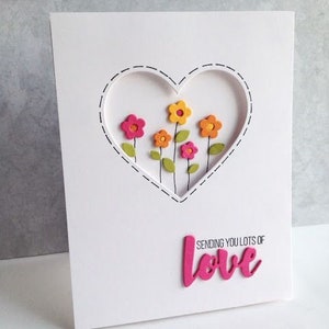 Puede incluir: Tarjeta de felicitaci&oacute;n blanca con un recorte en forma de coraz&oacute;n, con flores de papel de colores. La tarjeta incluye el texto "Sending you lots of love" en rosa.