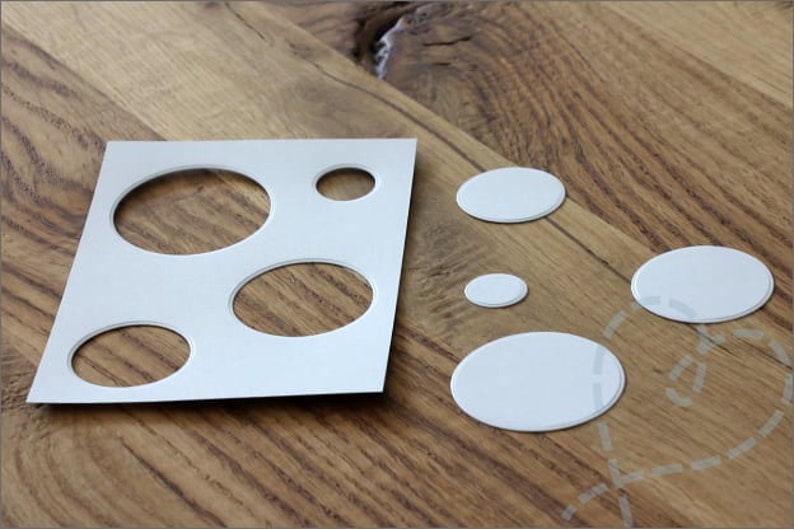 Puede incluir: Una plantilla de papel blanco con recortes ovalados y varias pegatinas ovaladas blancas se muestran sobre una superficie de madera. La plantilla tiene aberturas ovaladas de diferentes tama&ntilde;os. Las pegatinas son de diferentes tama&ntilde;os.