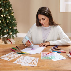Puede incluir: Una persona usa un bolígrafo azul y rojo para dibujar sobre papel blanco. Varios bolígrafos de colores, plantillas y una taza de té están sobre la mesa de madera. Un árbol de Navidad con adornos y regalos envueltos está en el fondo.
